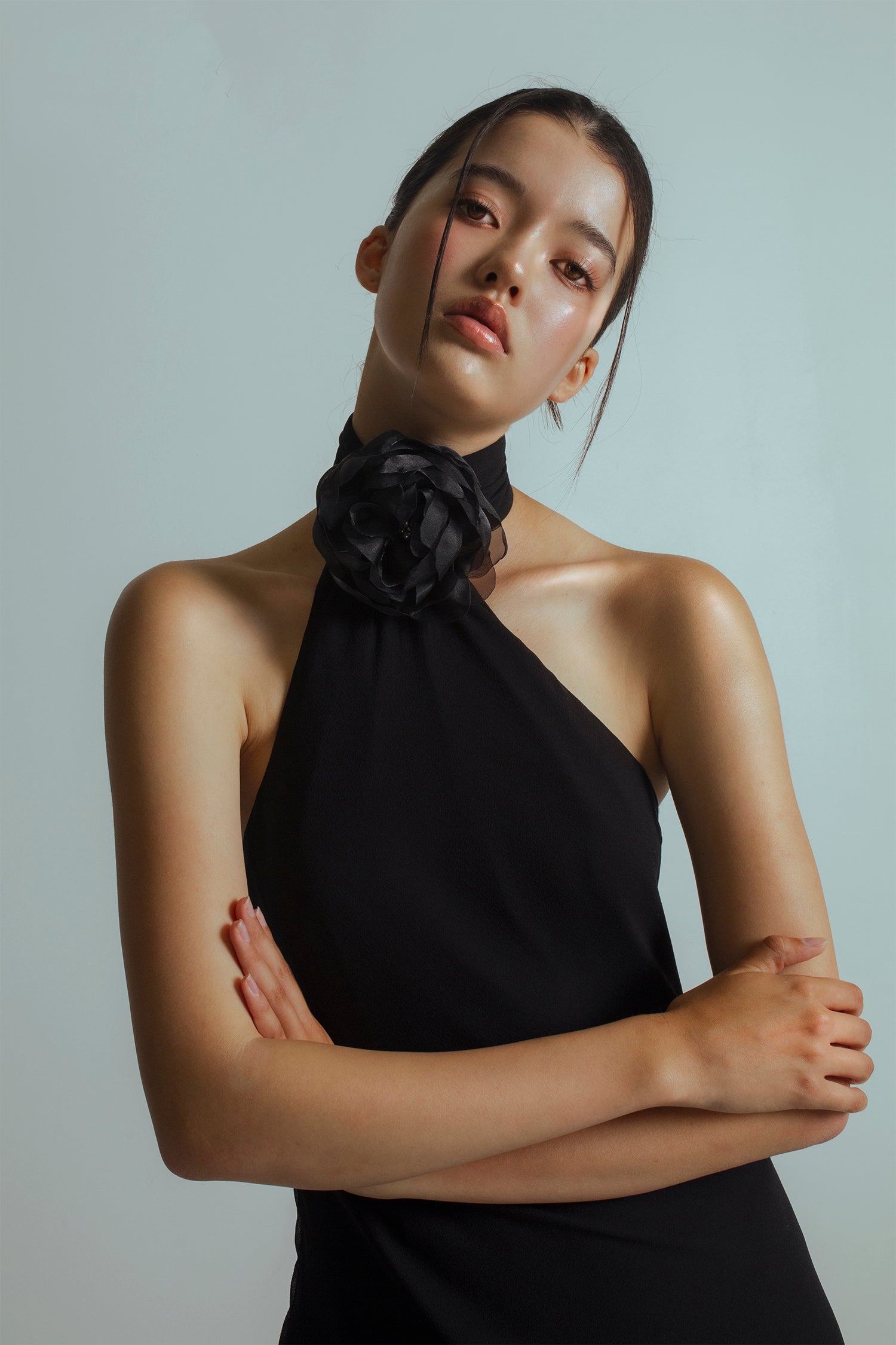 Half cropped image of model wearing a Black midi length dress with an asymmetrical neckline, detachable flower brooch at neckband and flowing ties at the back
