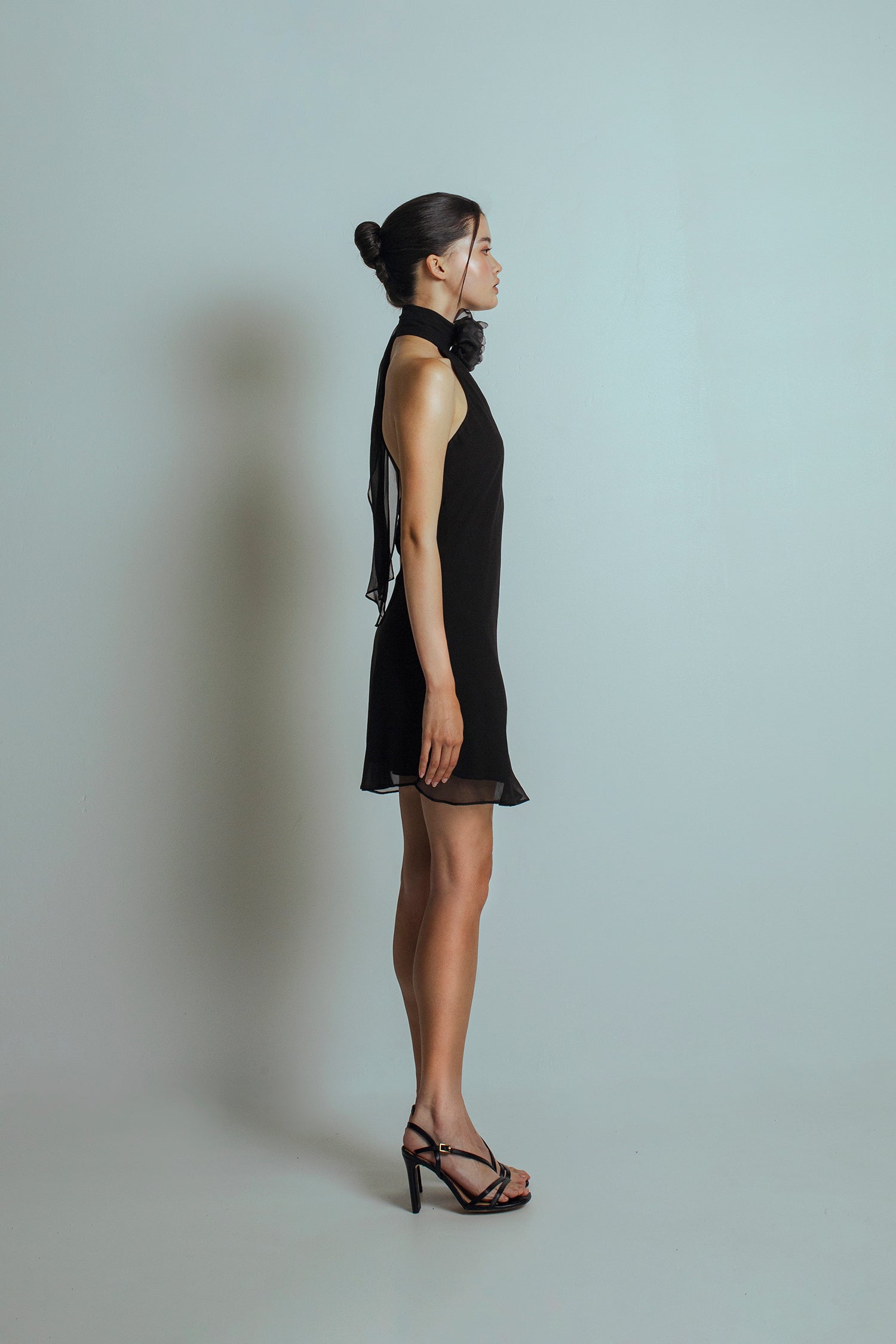 Side view of Black mini length dress with a detachable flower brooch at front neckband, bare back and flowing ties at the back neckband