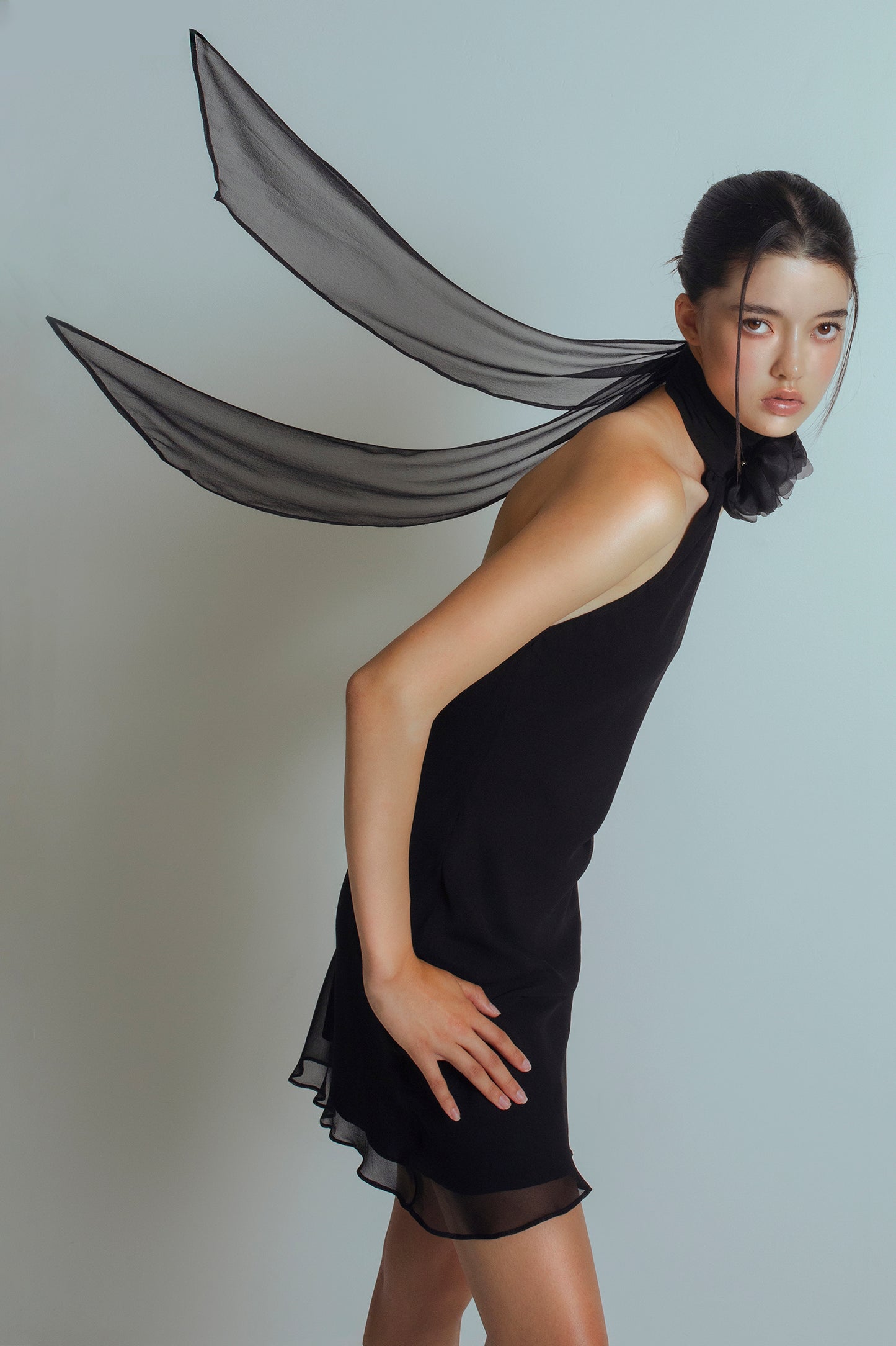 Half cropped image of model wearing a Black mini length dress with an asymmetrical neckline, detachable flower brooch at neckband and flowing ties at the back
